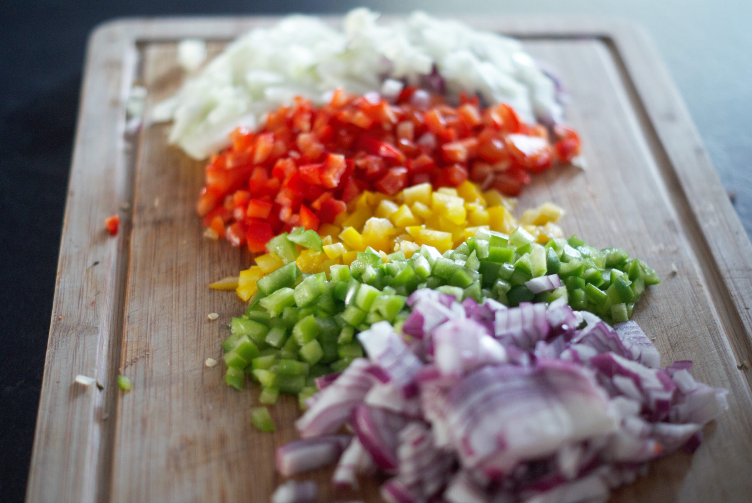 Chopped white onion, red pepper, yellow pepper, green pepper and red onion on a wooden chopping board